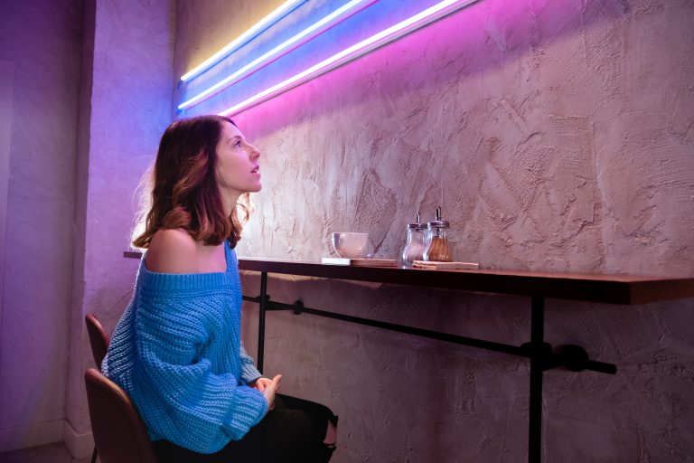 Woman sitting with cup on thin table against wall, looking up at neon lights.