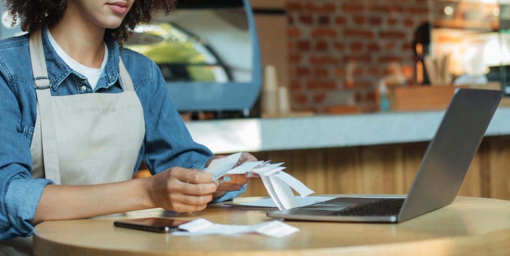 Shop owner checking receipts at table with laptop.