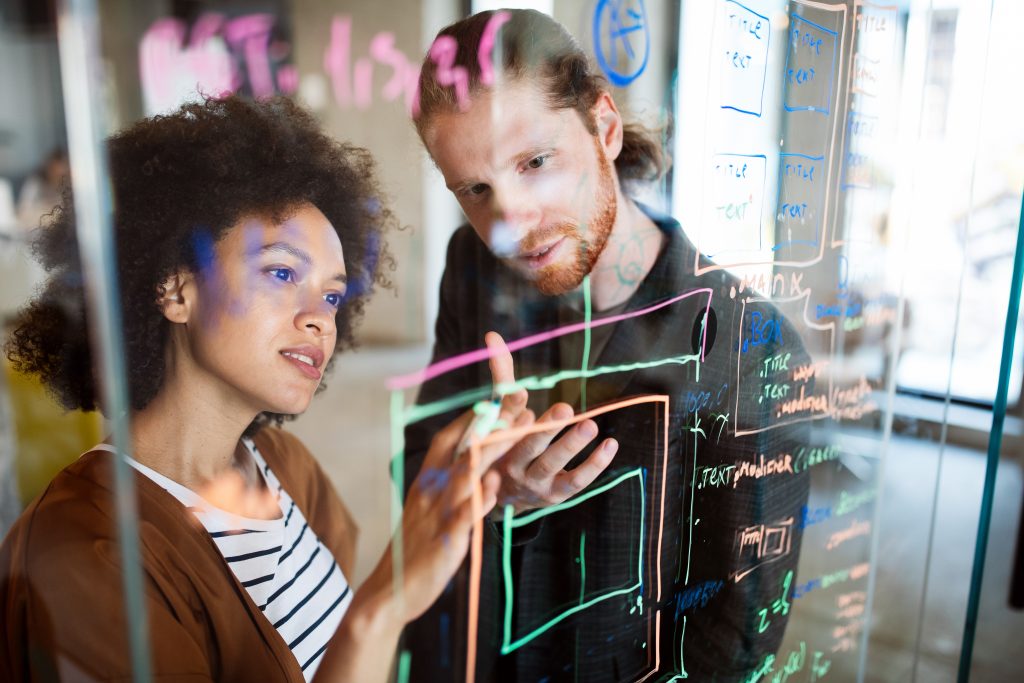 Two people discussing project ideas written on clear board.