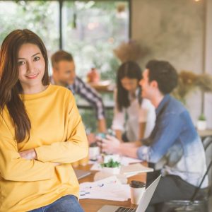 Person sitting on desk with arms crossed with people talking in background