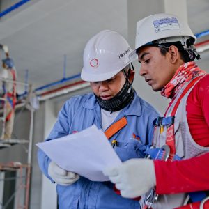 Two people in factory clothing with hard hats looking at paperwork.