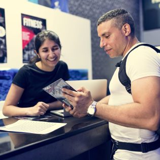 man wearing rucksack looking at flyer, person behind a counter
