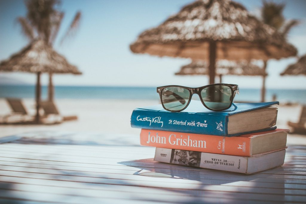 Three stacked books topped with a pair of sunglasses on a table at a tropical beach.