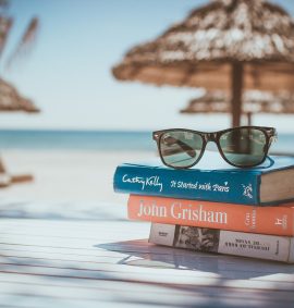 Three stacked books topped with a pair of sunglasses on a table at a tropical beach.