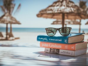 Three stacked books topped with a pair of sunglasses on a table at a tropical beach.