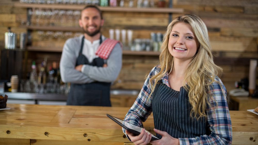 Smiling waitress holding digital tablet standing at bar, with barman behind out of focus.