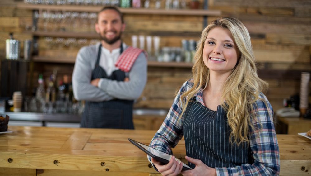 Smiling waitress holding digital tablet standing at bar, with barman behind out of focus.