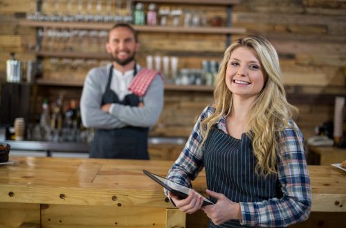 Smiling waitress holding digital tablet standing at bar, with barman behind out of focus.