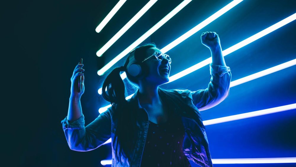 Woman wearing headphones listening and dancing in front of neon lights.