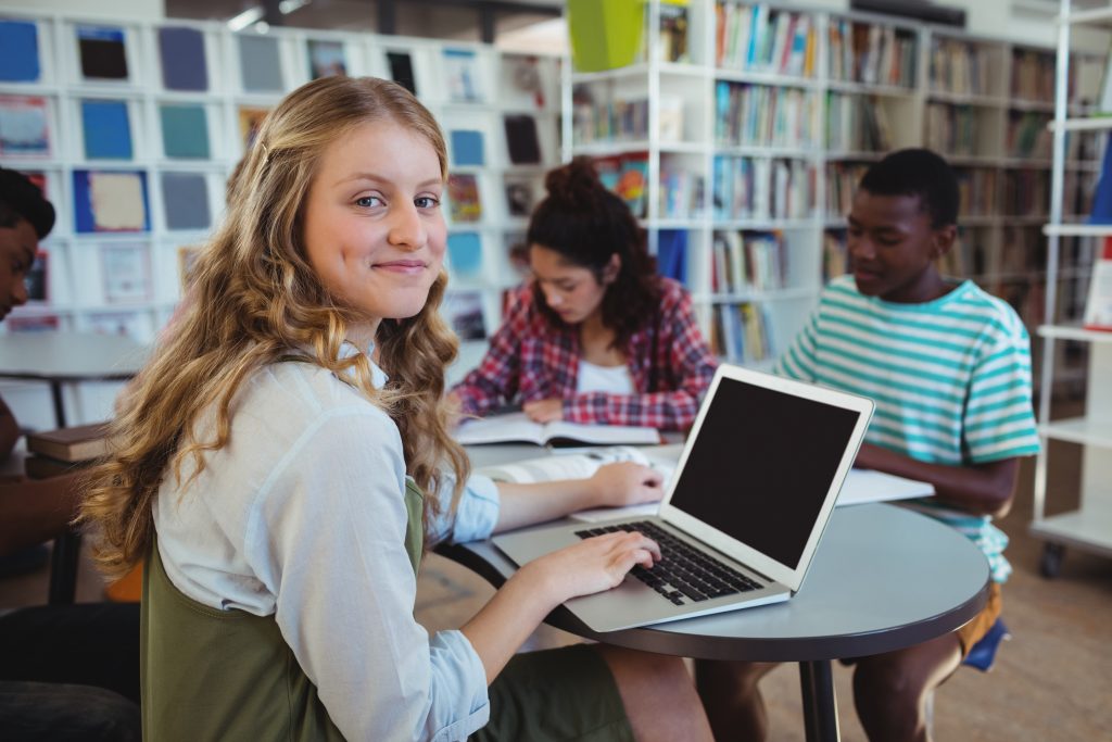 Sirl smiling while using laptop at table with other students in library.