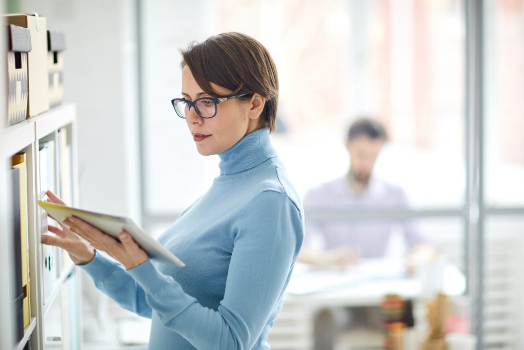 Woman checking digital tablet standing by filing cabinets.