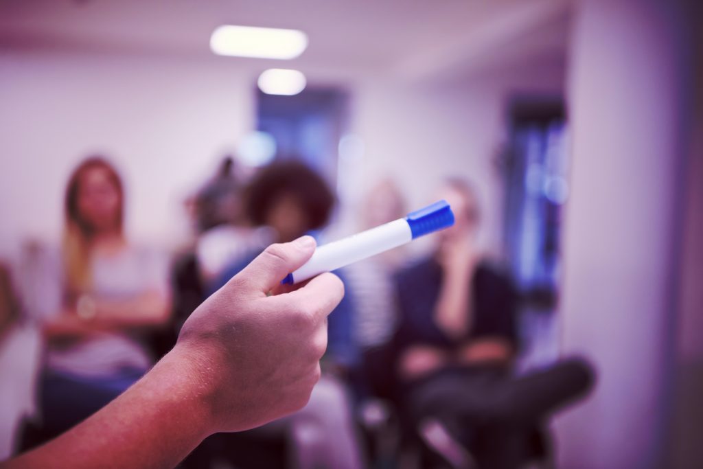 Close up of hand holding marker pointing with blurred students in background.