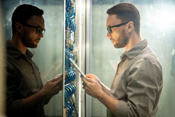 Man looking at digital tablet in front of network server rack.