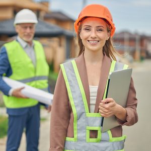 Woman wearing orange hard hat holding laptop at construction site.