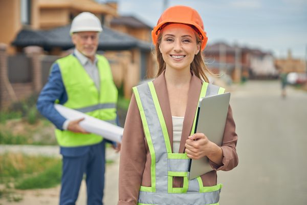 Woman wearing orange hard hat holding laptop at construction site.