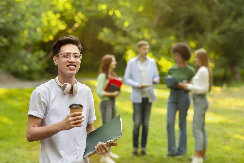 Learner holding a coffee and holding notebook with other learners in the background at a park