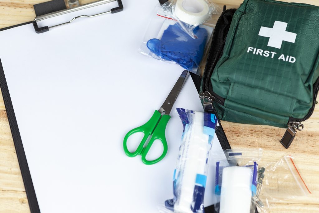 Pembrokeshire College First Aid at Work. Green first aid kit on a wooden table with a clipboard and a pair of scissors and some dressings