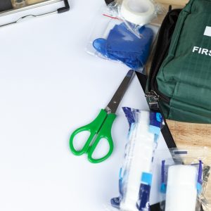 Pembrokeshire College First Aid at Work. Green first aid kit on a wooden table with a clipboard and a pair of scissors and some dressings