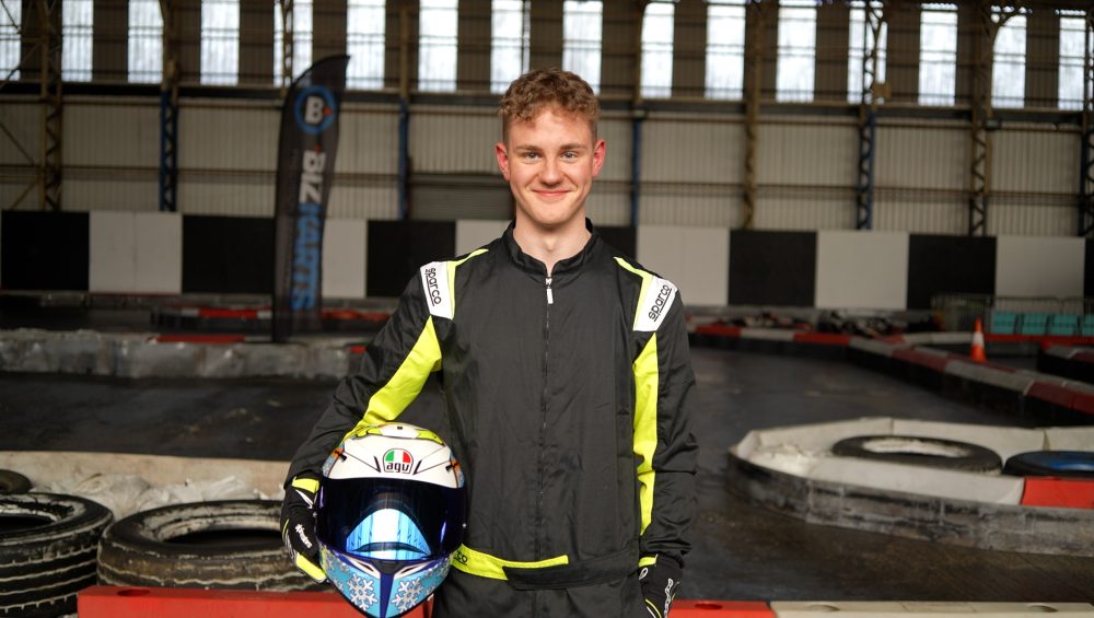 Reuben smiling at the camera whilst wearing black overalls and holding his white racing helmet. The go-kart track is in the background.