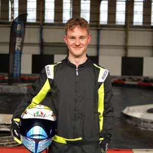 Reuben smiling at the camera whilst wearing black overalls and holding his white racing helmet. The go-kart track is in the background.