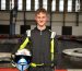 Reuben smiling at the camera whilst wearing black overalls and holding his white racing helmet. The go-kart track is in the background.