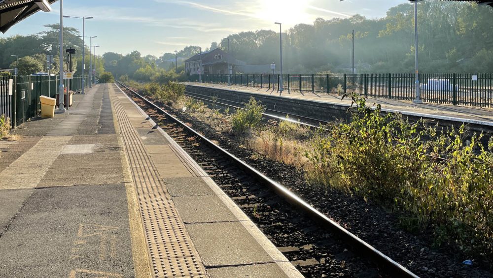 View along platform at train station, bridge and sun in background.