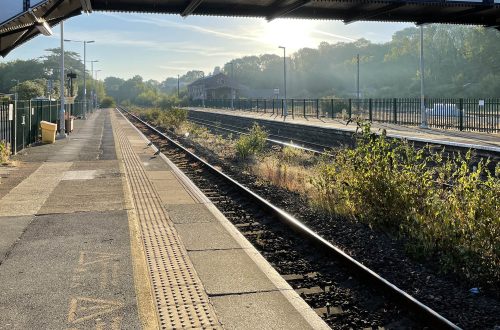 View along platform at train station, bridge and sun in background.