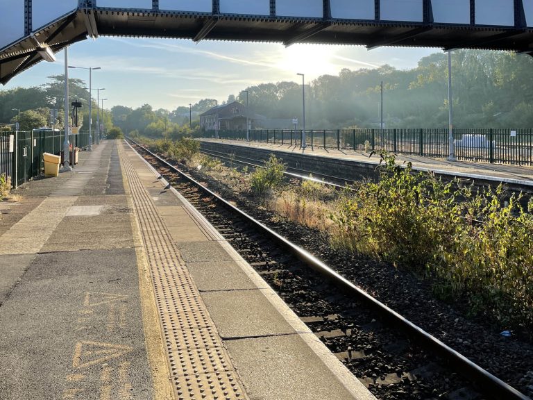 View along platform at train station, bridge and sun in background.