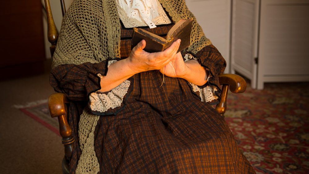 Older lady in traditional Welsh clothing on wooden rocking chair holding a book.
