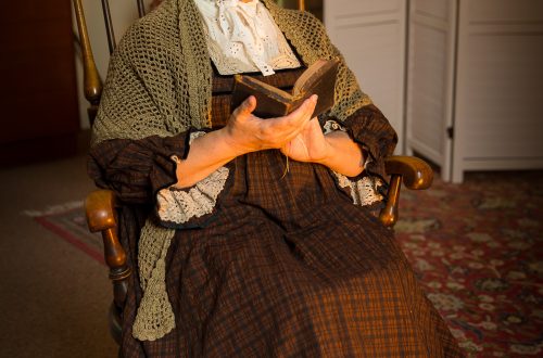 Older lady in traditional Welsh clothing on wooden rocking chair holding a book.