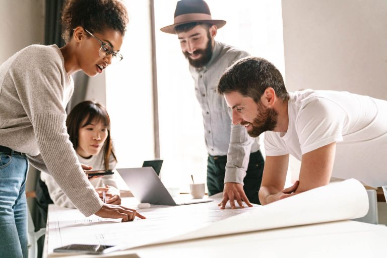 People leaning over and looking at large sheets of paper on a desk.