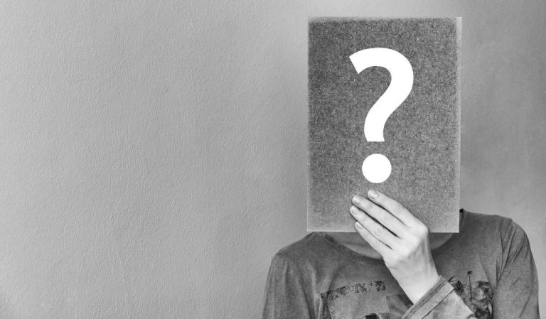 Black and white image a person holding a board in front of face with a large question mark on it.