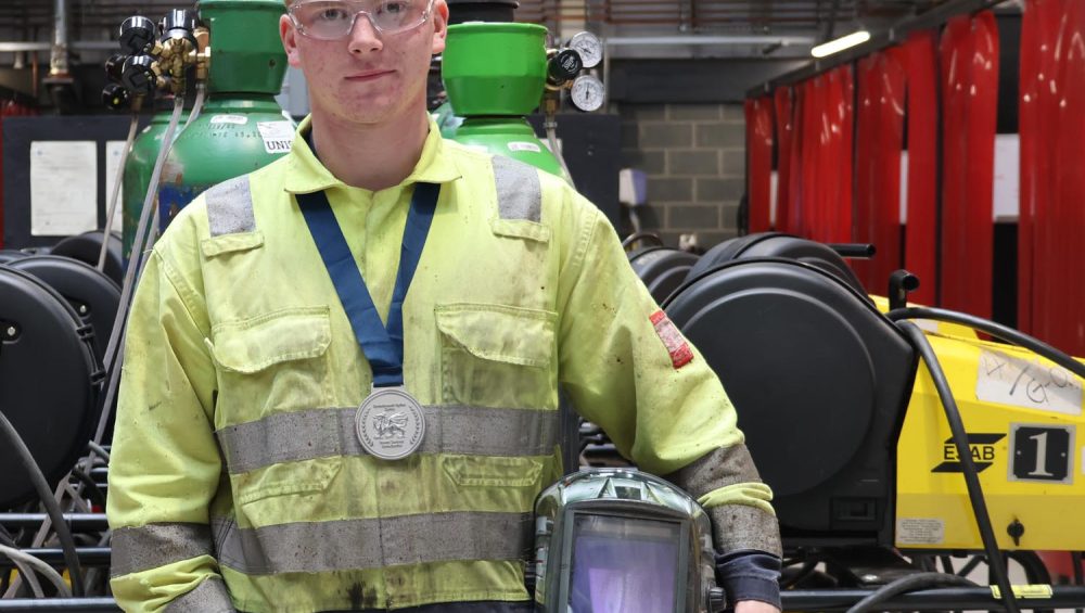 Leaner Luke wearing medal and holding welding helmet in workshop.