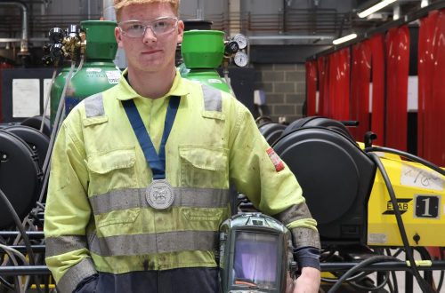 Leaner Luke wearing medal and holding welding helmet in workshop.