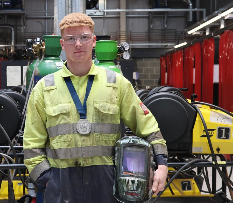 Leaner Luke wearing medal and holding welding helmet in workshop.