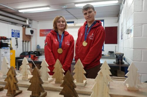 Two students wearing red Worldskills hoodies in workshop with wooden trees in foreground