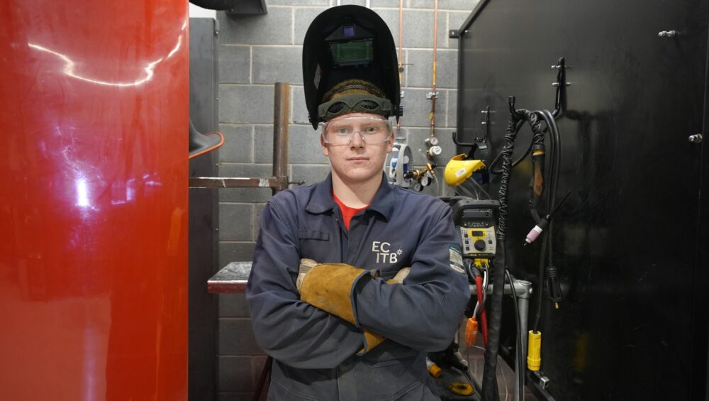 Luke with arms folded located in a welding booth wearing his visor