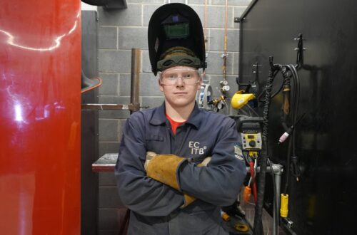 Luke with arms folded located in a welding booth wearing his visor