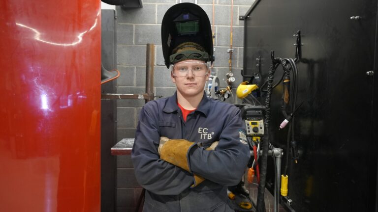 Luke with arms folded located in a welding booth wearing his visor