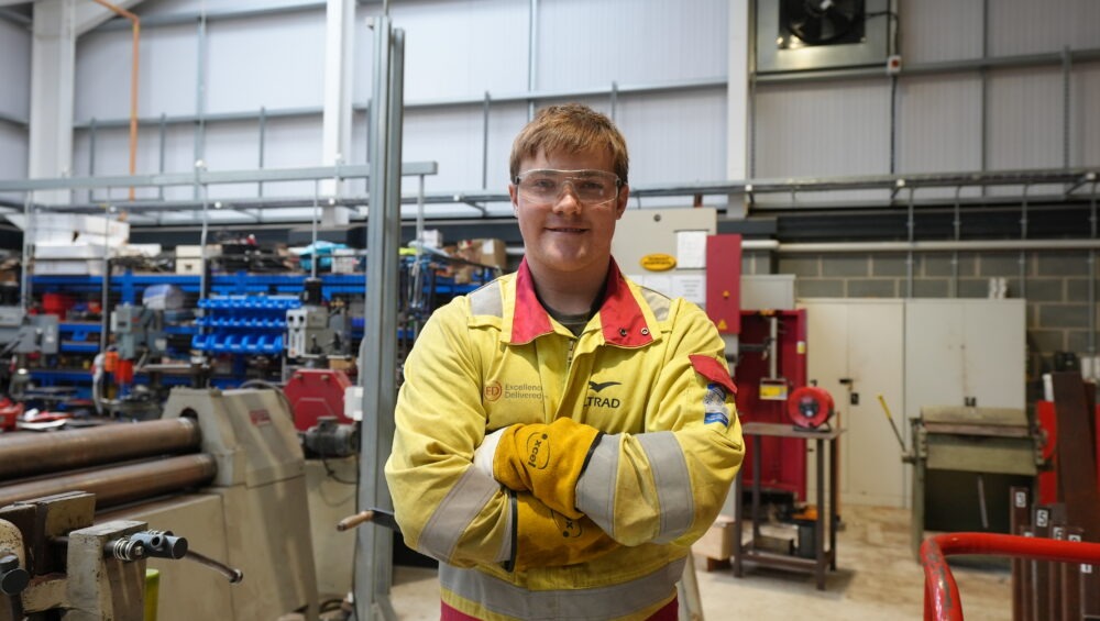 Student wearing yellow overalls with arms crossed in a workshop.