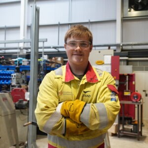 Student wearing yellow overalls with arms crossed in a workshop.