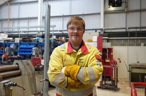 Student wearing yellow overalls with arms crossed in a workshop.