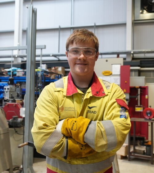 Student wearing yellow overalls with arms crossed in a workshop.