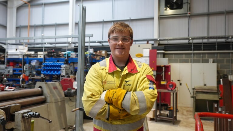 Student wearing yellow overalls with arms crossed in a workshop.