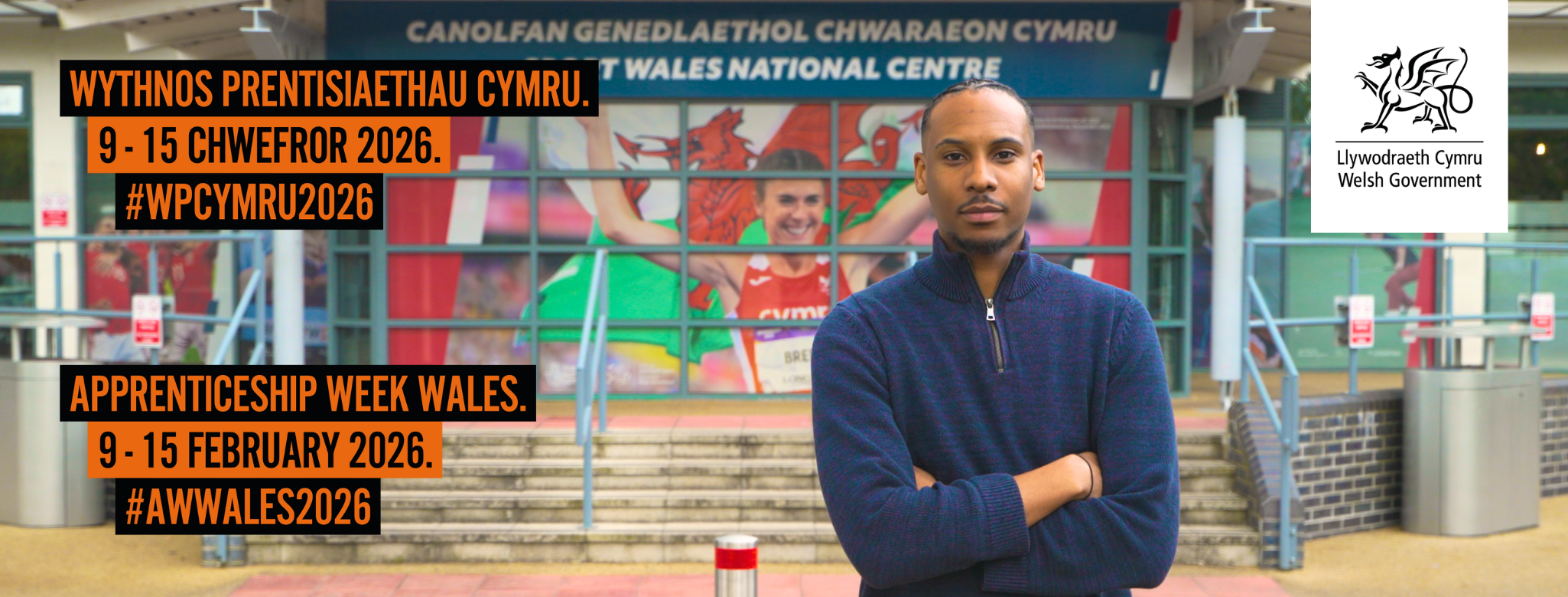 Man standing with arms crossed outside stadium. Text says: Apprenticeship Week Wales 9 - 15 February 2026.