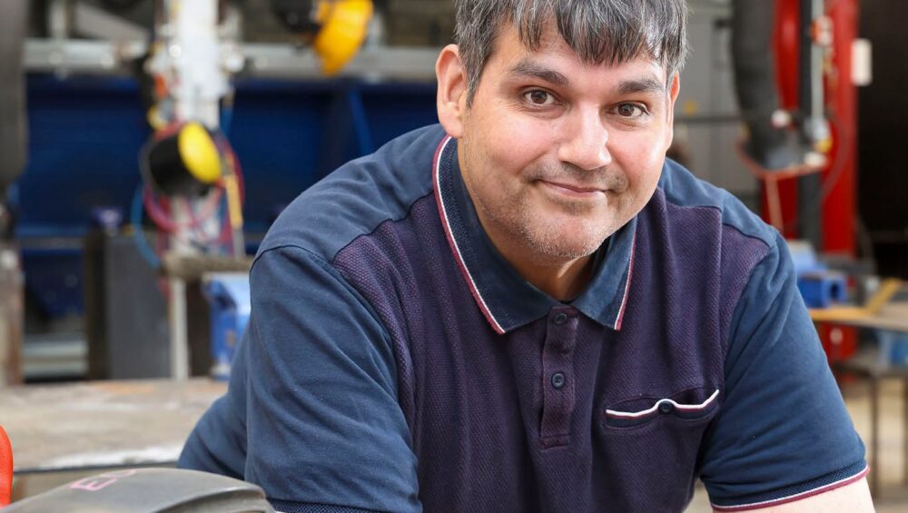 Member of staff leaning on workshop bench next to welding helmet.