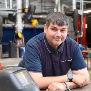 Member of staff leaning on workshop bench next to welding helmet.