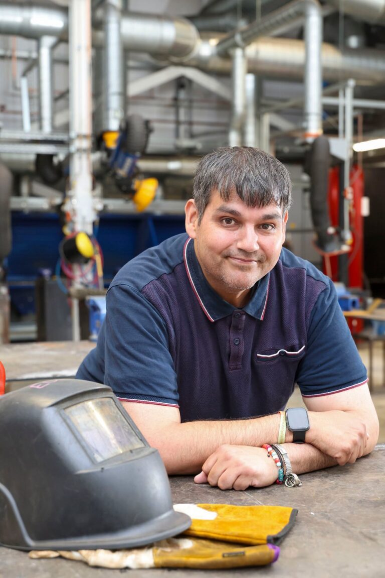 Member of staff leaning on workshop bench next to welding helmet.