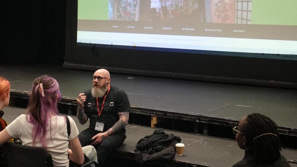Phil sitting on stage, with images of tattoos in background and learners seated in theatre.