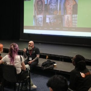 Phil sitting on stage, with images of tattoos in background and learners seated in theatre.